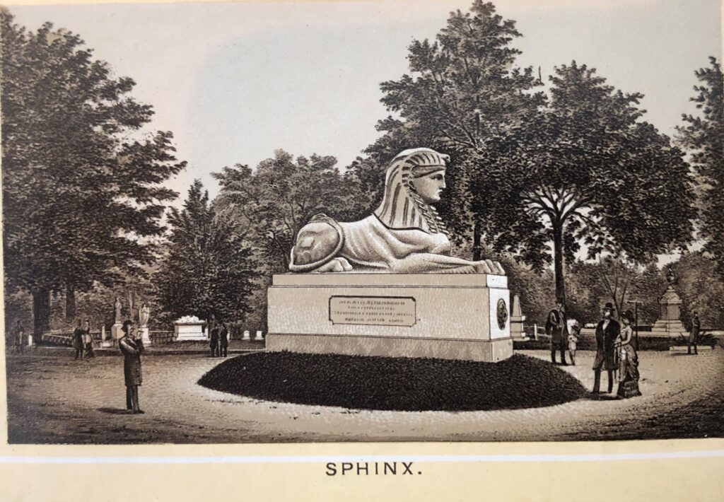 Sepia view of an ornamental cemetery landscape in the 19th century with visitors on a road or clearing looking up at a large statue of an Egyptian sphinx sitting on a large pedestal on a small hill of grass. Trees, monuments and other strolled are in the background. Below text reads “Sphinx.”