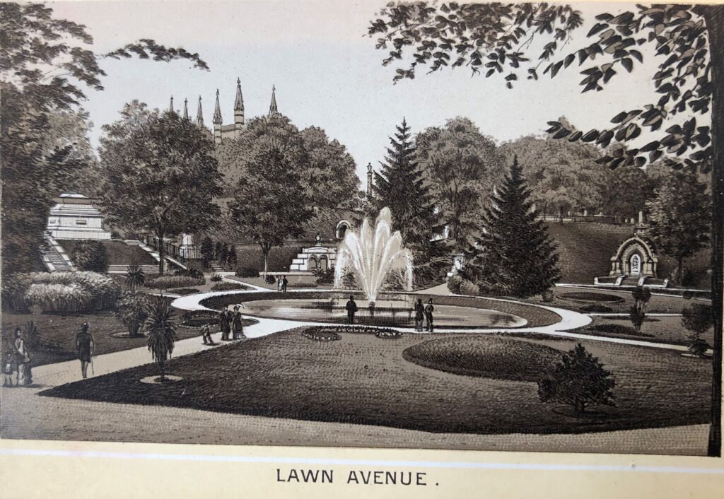 Sepia view of an ornamental 19th-century cemetery landscape with a central circular pond and fountain, surrounded by curbing, grass lawns, palms, deciduous trees, large monuments and in the background, the spires of a small chapel on a hill. Below text reads “Lawn Avenue.”