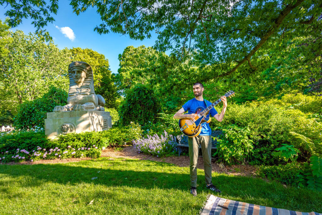 Ira Klein holding a guitar standing on the lawn next to the Sphinx Memorial