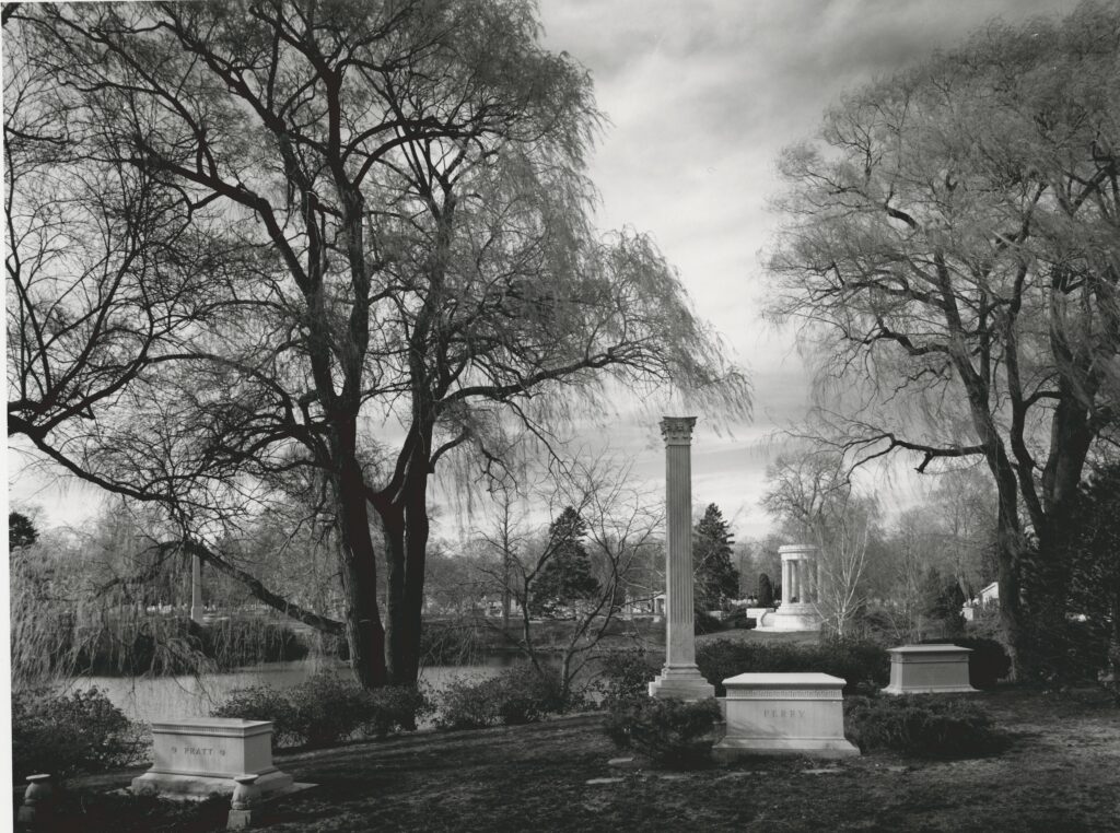 Black-and-white photograph of a cemetery landscape with memorials. large trees, and plantings around a lake.