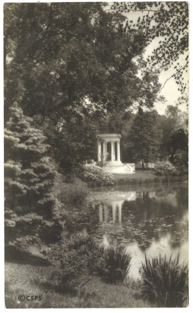 Black-and-white photograph of the shore of a lake with trees and plants, showing a white circular memorial with columns on the far side. The memorial is reflected in the water.